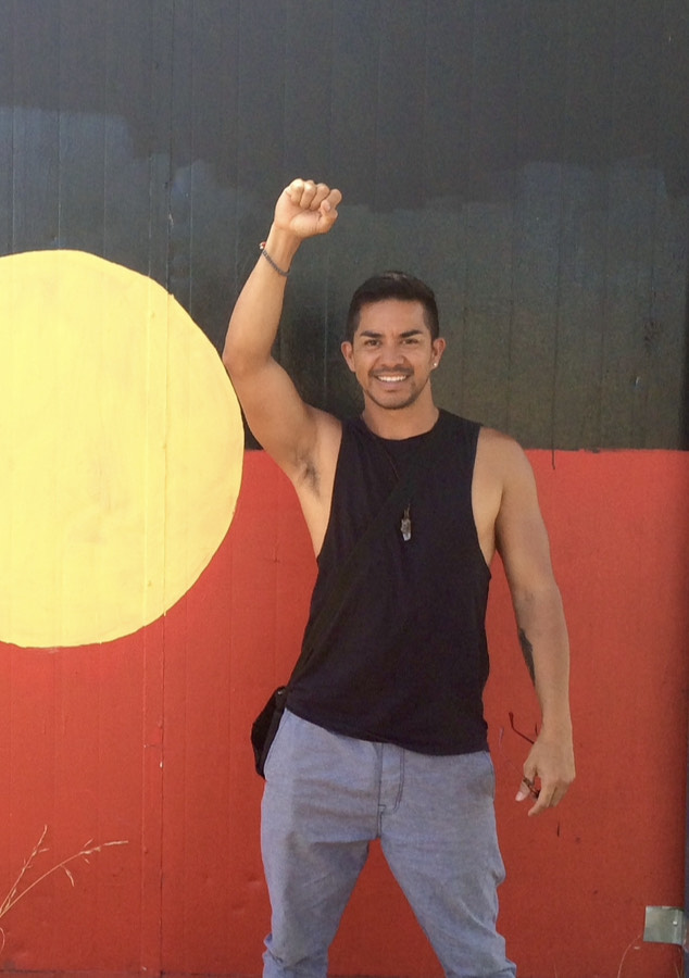 Sermsah Bin Saad stands in front of a wall painted to be the Australian Aboriginal flag, with one arm raised and a smile.