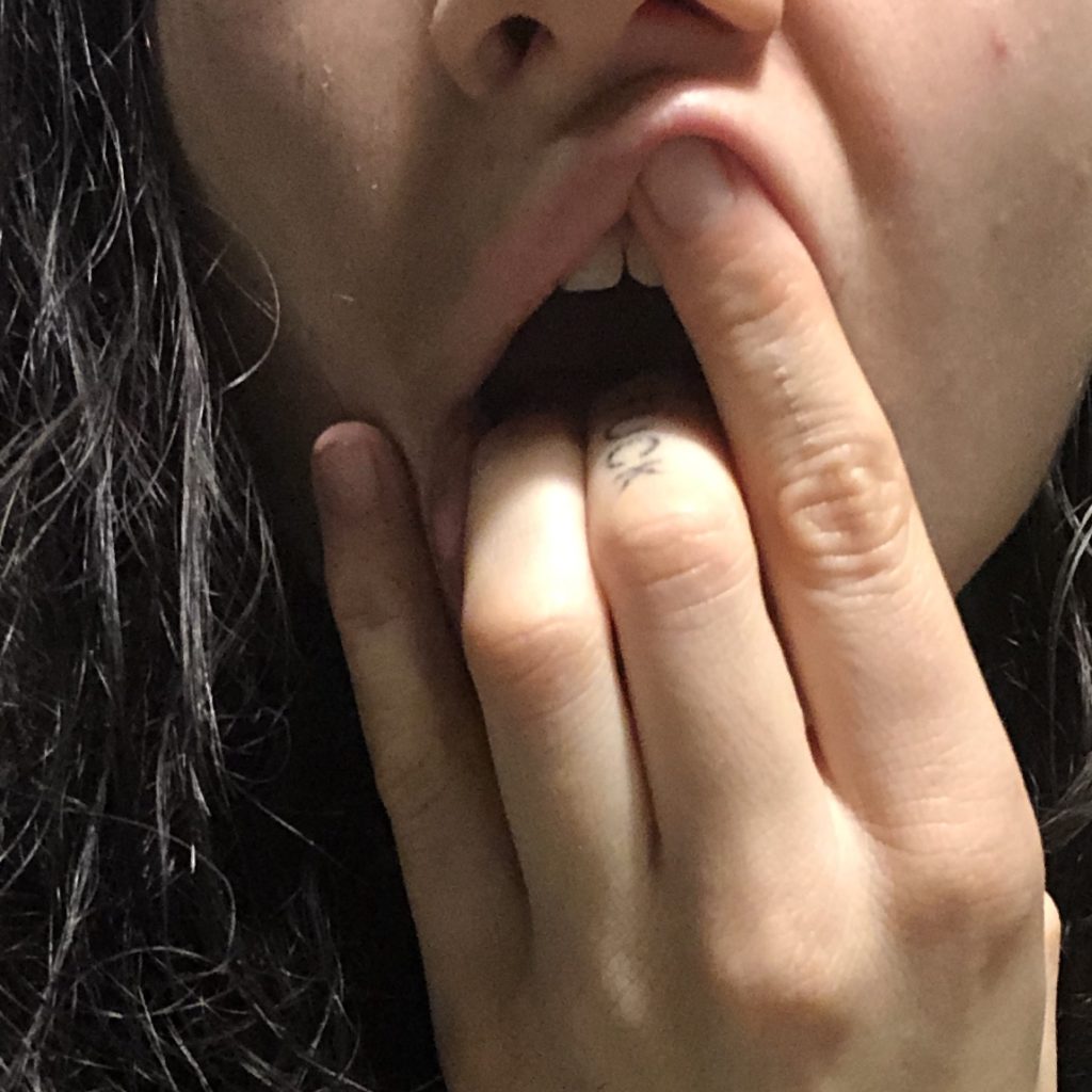A close up image of Nayook's fingers and mouth. They are pushing their fingers into their open mouth holding down their tongue and pushing up their top lip. We can see their nostrils and the rest of the frame is filled with their dark curly hair.