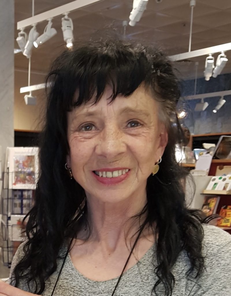 Jeanine Leane is standing ina book store smiling at the camera. They have long black curly hair, that goes past their shoulders and with a curled front fringe. They have red lipstick on and small dangly earrings.