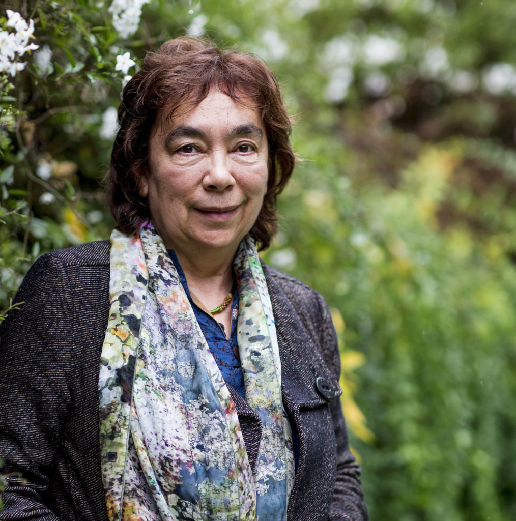 Alexis Wright is standing outside infront of a lush green garden. She is looking at the camera, wearing a grey coat and a floral scarf draped over her shoulders. She has chin lengths nutty brown hair.