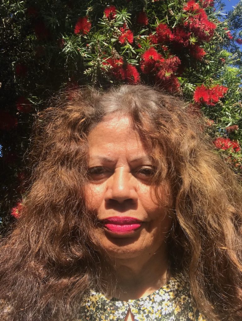 Aunty Bev Murray is outside infront of a banksia tree. The red flowers can be scene behind her. It is a very sunny day. Aunty Bev has long wavy brown hair that goes past her shoulders. SHe is wearing a hot pink lipstick, and a collared floral shirt.