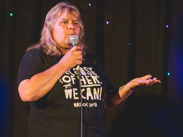 Aunty Denise McGuinness is standing on stage in front of a black curtain with fairylights. She is holding a microphone with one hand and gesturing out with the other. She is wearing a black t-shirt with white text. She has long blonde hair and a front fringe.