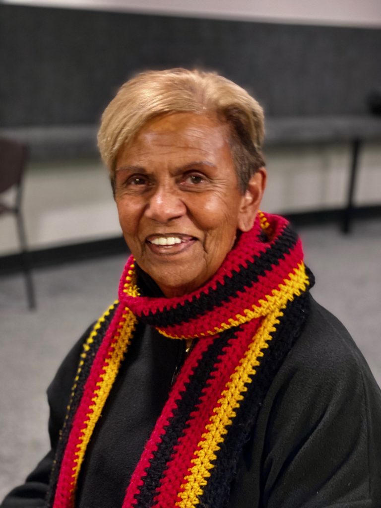 Aunty Pam Pedersen's headshot. She is looking into the camera, smiling. She has a knitted black, yellow and red scarf wrapped around her next, and wearing a black top. She has short light hair.