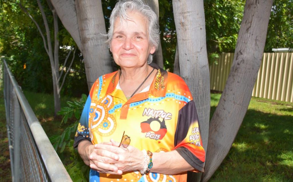 Aunty Pattie Lees is outside standing under a tree and next to a metal fence. Her hands are clasped together with her glasses held in her hands also. She is smiling looking at the camera, and has short and wavy grey hair. SHe is wearing the NAIDOC 2015 polo shirt, that is very colourful and has many Aboriginal themed graphics, such as the aboriginal flag,and traditional painting.