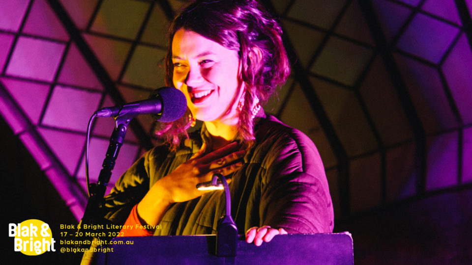 Jazz Money is standing at a podium under purple and yellow lights. She has one hand on the lecturne, and their other hand on their chest. They are smiling and looking to the left of the camera. They have long dark hair that is tied back ,with pieces falling out, fair complexion and wearing a dark top with long sleeves.