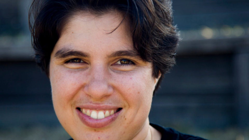 A close up photo of Ellen van Neerven's face. They are outside on a sunny day, infront of a building that is out of focus in the background. They have a fair skin complextion, and short hair that frames their face and reaches their eyebrows. They are smiling widely at the camera.