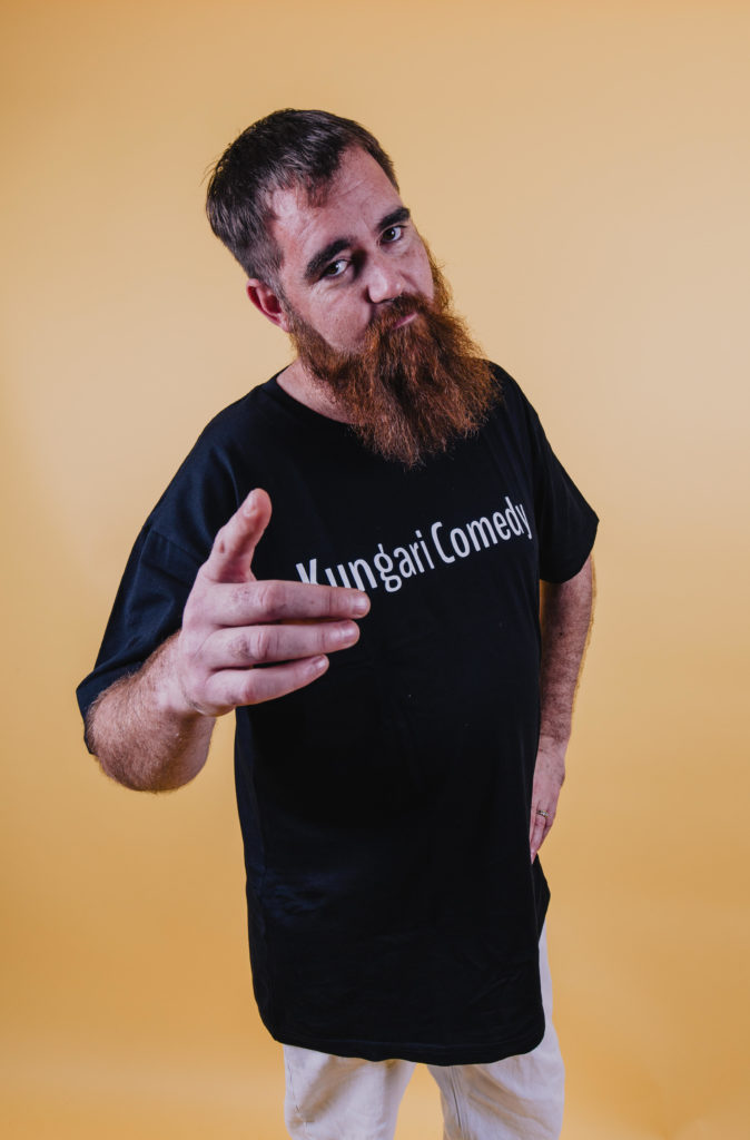 Ben Moodie is standing in front of a light orange background looking directly at the camera. His right hand is pointing directly to the camera, and his left hand is on his hip. He is wearing a black t-shirt with white text across his chest that reads 'Kungari Comedy'. He has a fair complexion with a red blush, short brown hair on his head, and a long orange toned beard.