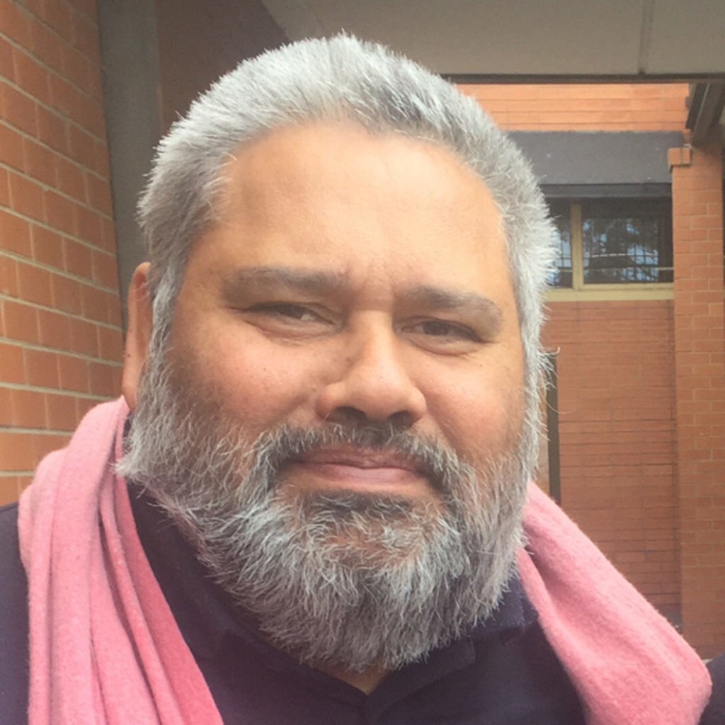 Glenn Shea headshot showing his face and shoulders. He is wearing a black top with a pink scarf draped around his neck. He is looking directly at the camera, and has short but styled grey hair and a beard.