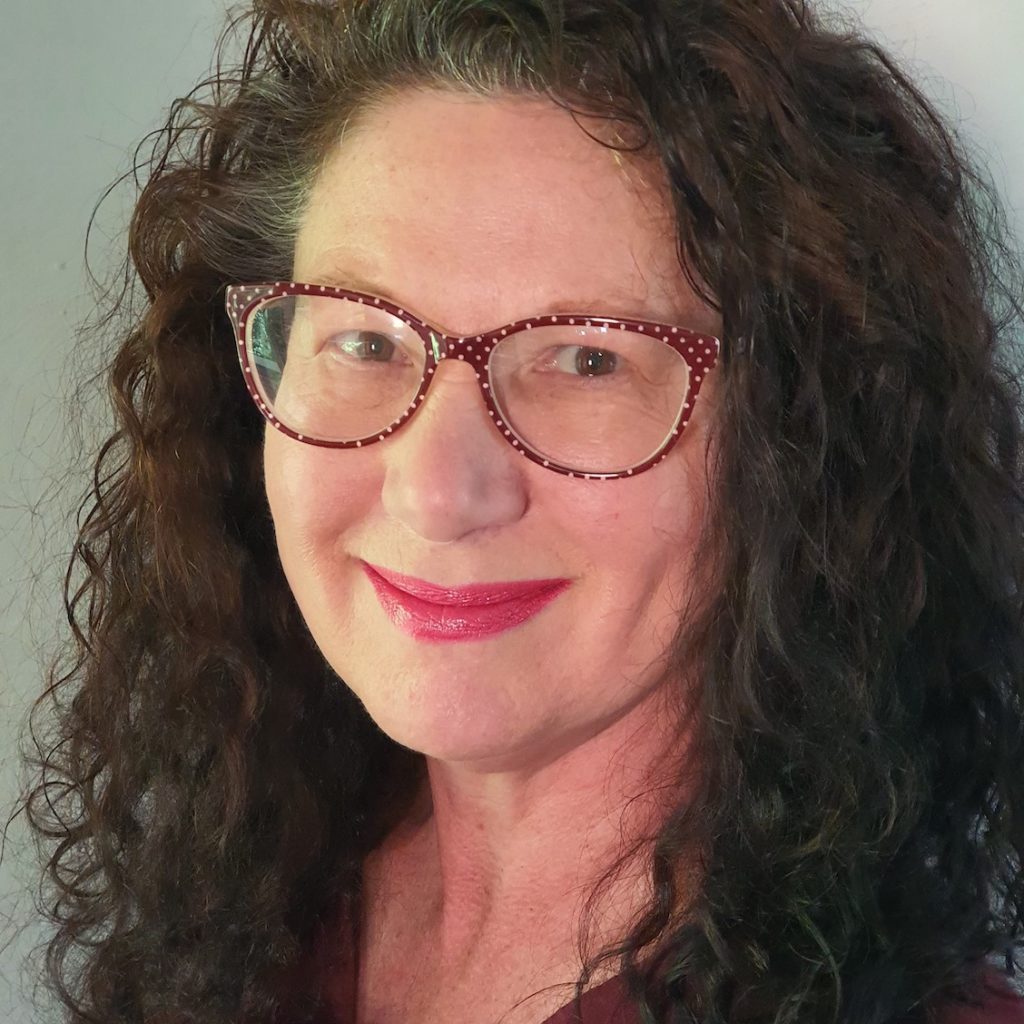 Jane Harrison's headshot. It is a close up picutre of Jane's face. She has long, curly dark brown hair which frames her face and rests on her shoulders. She is wearing glasses with red frames that have white polka dots. SHe is smiling at the camera and wearing red lipstick.