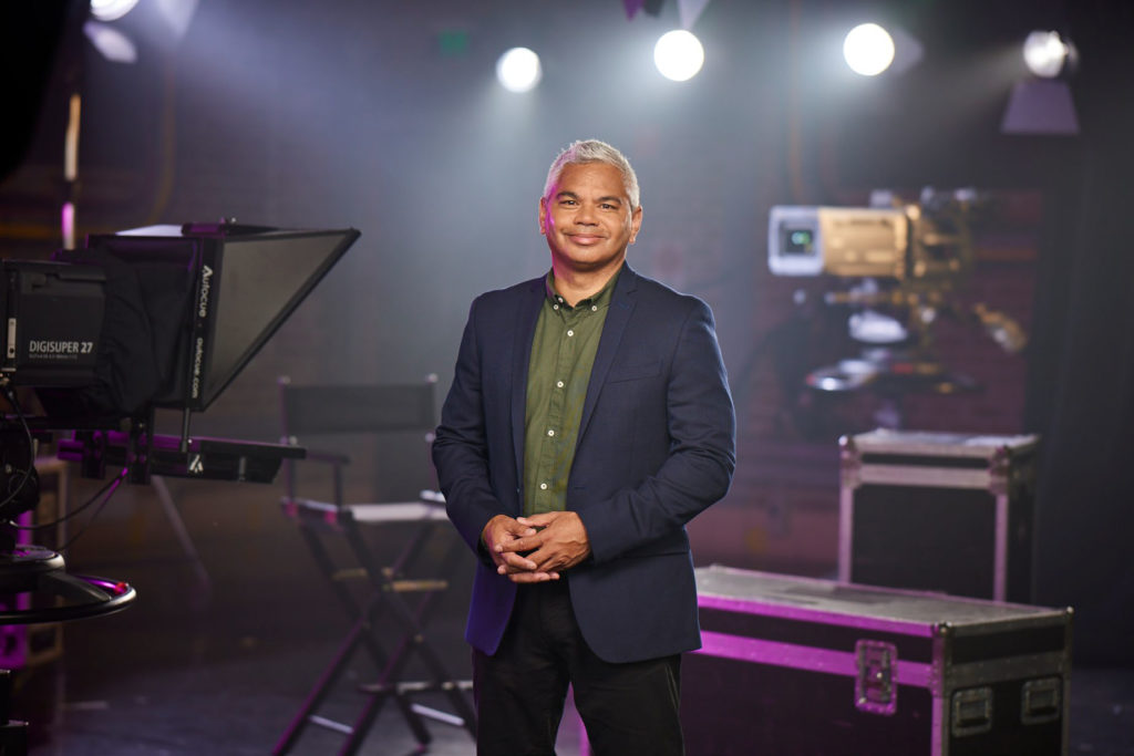 John Paul Jenke stands in a blue suit with a green shirt on a tv set. There are bright lights behind him and camera equioment. He stands with his hands clasps, smiling and looking directly at the camera.