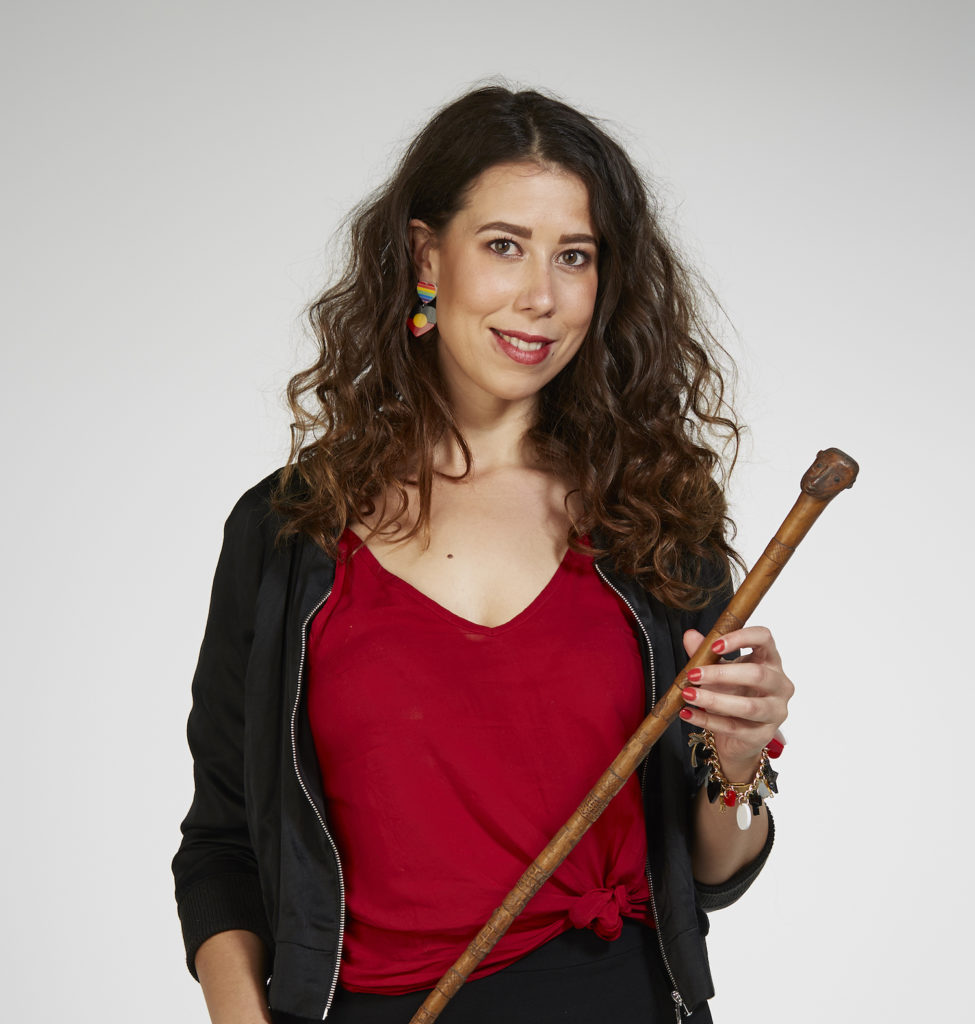Kimberley Moulton's headshot. She is standing infront of a white wall, holding a traditional instrument that is a stick with intricate engravings along it, across her body. He has long brown curly hair, red lipstick, and red nail polish. She is wearing a black sports jacket with a red, v-neck t-shirt that is tied in a not to oneside. She has graphic earings that are made up of two lovehears ontop of each other. The top with rainbow stripes, the bottom with the Aboriginal flag.