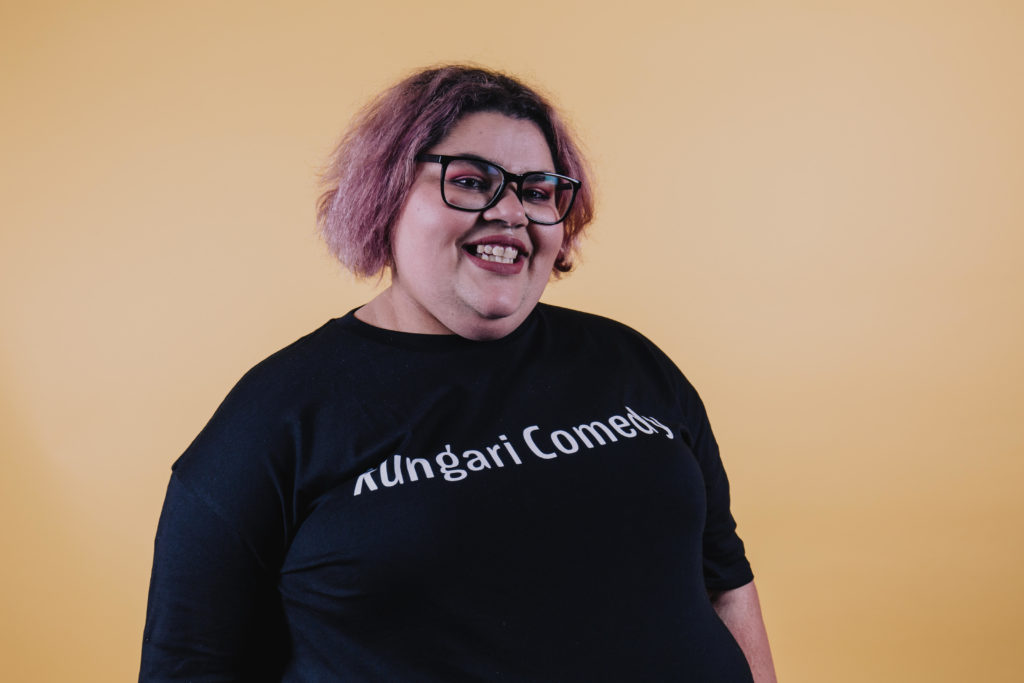 Kimmie Lovegrove is standing in front of a light organise background. She is smiling at the camera, wearing plum coloured eyeshadow and lipstick and wearing a black t-shirt with white text across her chest that says 'Kungari Comedy'. She has black framed glasses, short hair that reaches just past her ears that is died pink at the ends and black at the roots.