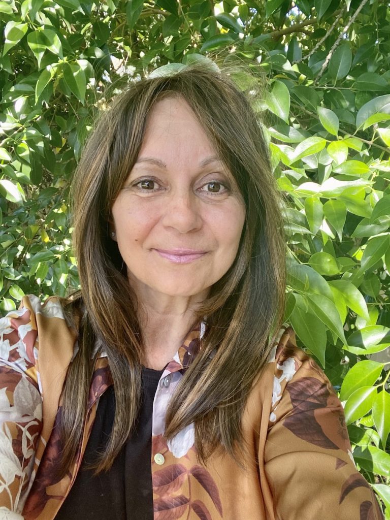 Leonie Drummond is standing infront of a very leafy, bright green tree on a sunny day. She has long straing hair, brown with some highlights through it. She is smiling at the camera, wearing a satin pink lipstick. She is wearing a silk shirt that is brown and gold with floral details.