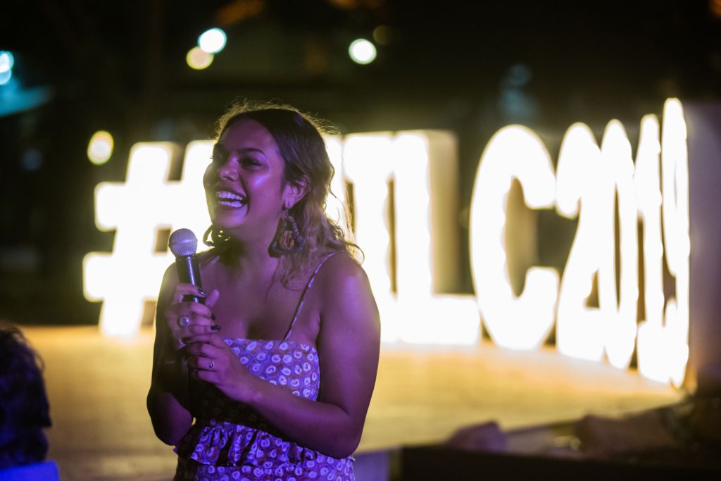 Merinda is holding a microphone, laughing and looking just left of the camera. It is night time, and she is standing in front of a large lit-up sign made out of letters. She has long hair, dark at the roots and lighter at the ends, wearing a dress.