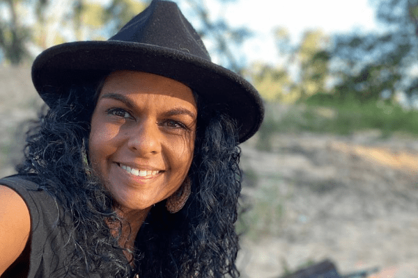 A selfie of Teela Reid where she is outside in nature. She is wearing a black, widebrim hat that causes some slight shadow to fall onto her face. She has long,black curly hair and is smiling directly at the camera.