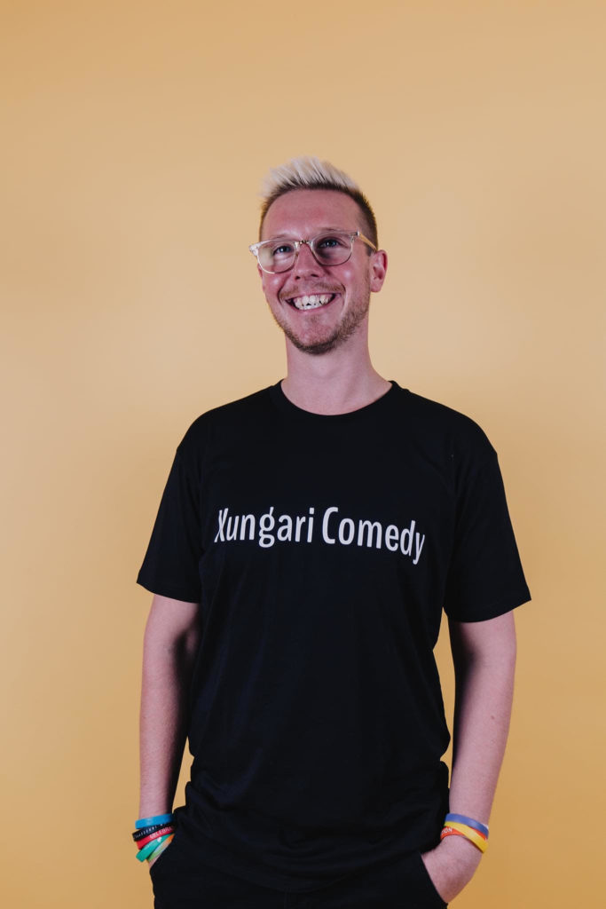 Tommy Pemberton is standing infront of a light orange backfround smiling just past the camera. He is wearing a black t-shirt that has white text across his chest that says 'Kungari Comedy'. He hair is shaved at the sides, but spiked upwards on top and died blonde. He has clear framed glasses, and 3 differently coloured plastic bracelets on each wrist.