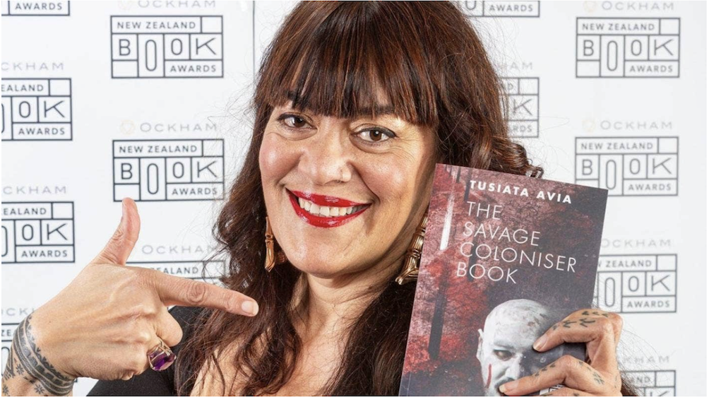 Tusiata Avia standing infront of a black and white promo wall, holding up her book 'The Savage Coloniser Book' with one hand, and putting her finger towards the book with another. She is smiling, looking at the camera, wearing red lipstick, and earrings. She has long reddish-brown hair, that is curled with a front fringe. She has many tattoos shown on her wrists and fingers.