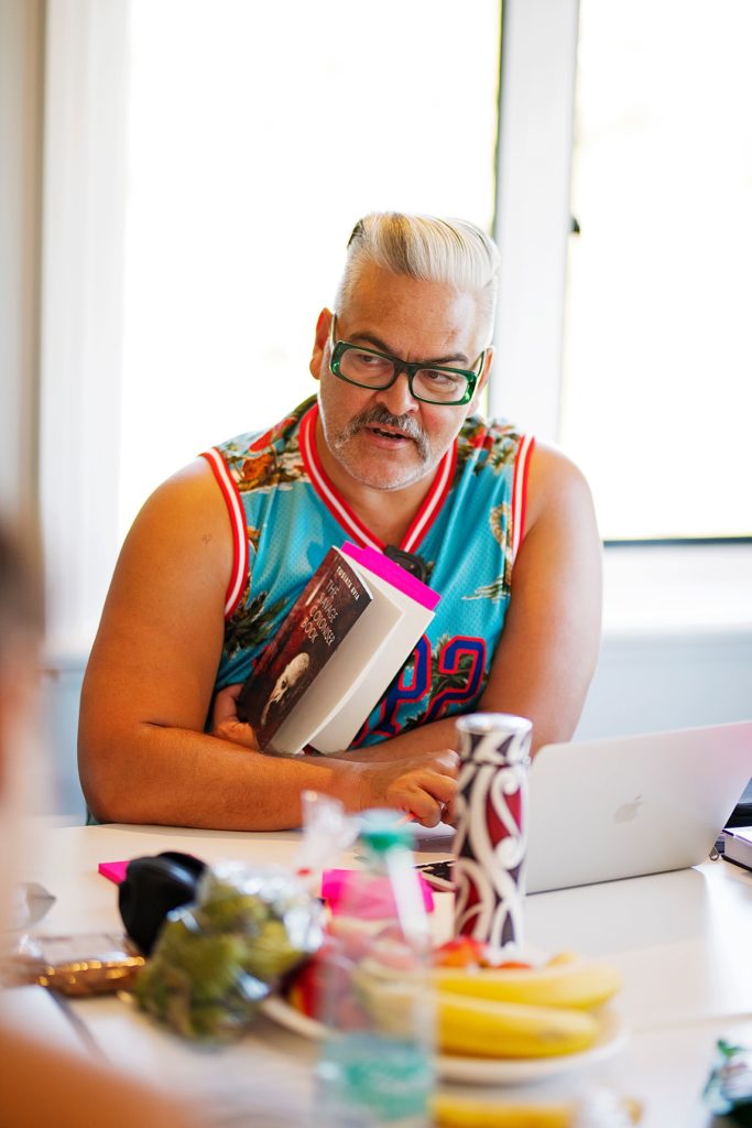 Victor Rodger is sitting at a table in an office. There are other people around the table that are out of focus. He is leaning over his laptop and holding a book with pink post-it notes sticking out. He is loioking to the left of the camera. He has combed back grey hair, green framed glasses, and a colourful top in the style of a basketball Guernsey.