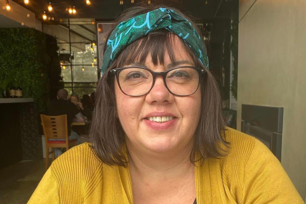 Dr Crystal Mckinnon stares at the camera smiling. It shows her shoulders and face, she is wearing a yellow top, black framed glasses and a blue and green headband. She has short brown hair styled in a bob with a front fringe.