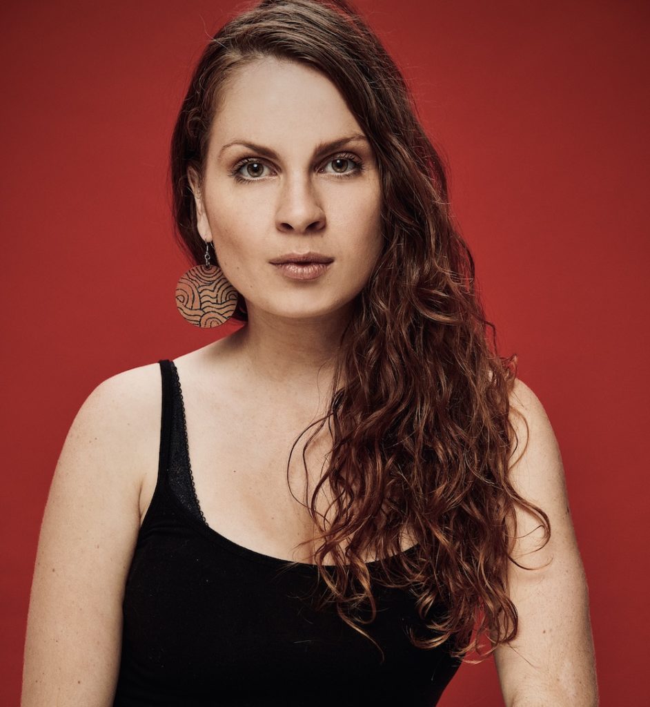 Headshot of Monica Karo. Monica is squatting infront of a red background. We can see from their knees up. They have long, wavy, brown hair that reaches their chest, and is pulled aside to one side. They have a fair skin tone, and are wearing a black tank top, dark jeans and a belt with silver metals. They have large circle dangling earring and are staring straight into the camera.