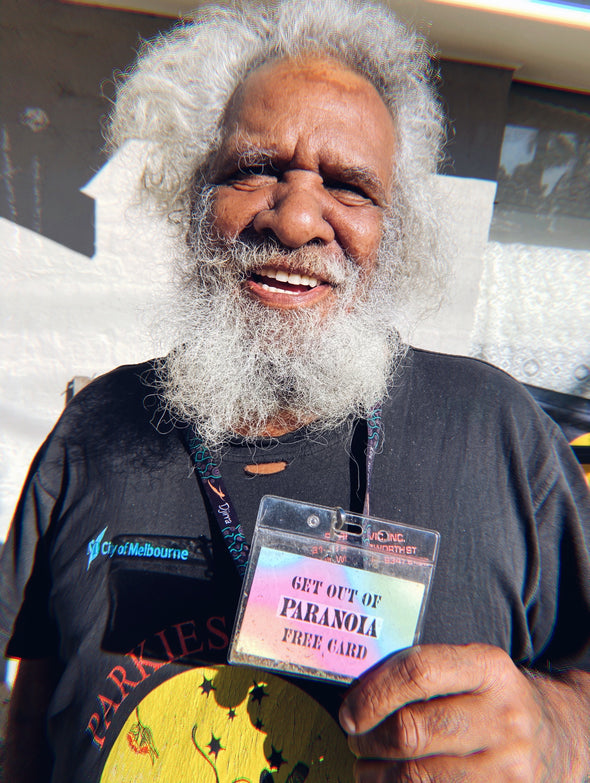 Uncle Talgium Edwards is looking at the camera holding up lanyard that reads "Get out of Paranoia free pass" He has curly greay hair and beard that frames his face like a mane. The sun is lighting his face, he is outdoors, and there is a cement building behind him