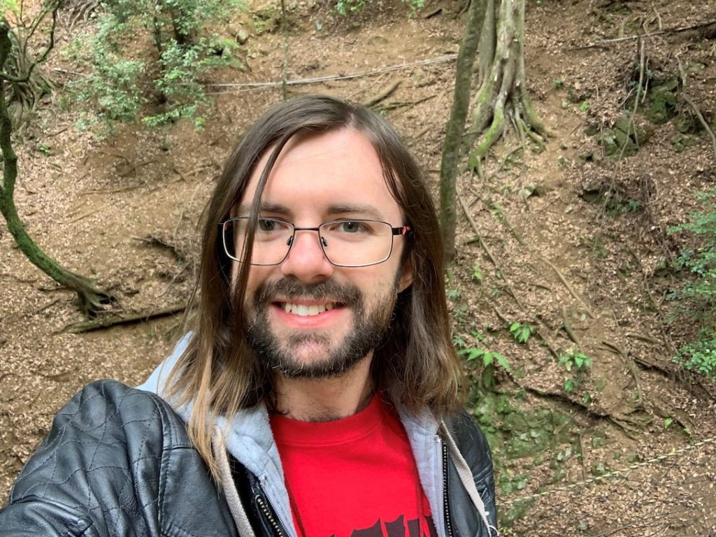 Beau Windon is shown from his chest up, smiling and looking at the camera. He has a fair complextion, straight, shoulder length hair that is brown with blonde ends. He is wearing a leather-looking jacket, with a grey hoodie underneath. Both are open to show a read t-shirt. He has square, thin framed glasses, and a short beard. He is standing outside, where mossy tree roots, and dirt can be seen behind him.