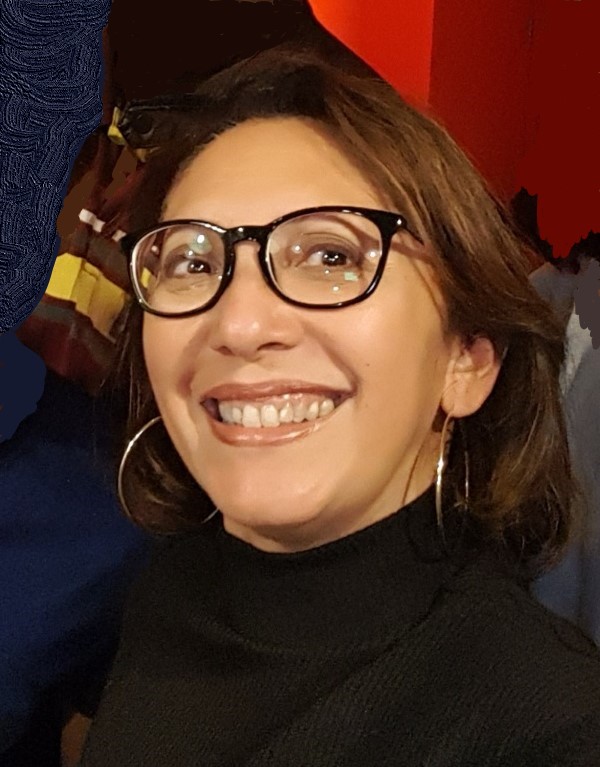 Lenora is wearing blackrimmed square glasses, has light brown short hair that sits behind her ears. She has large, thin silver hoops in her ears, and a black turtle neck. She is smiling just past the camera.