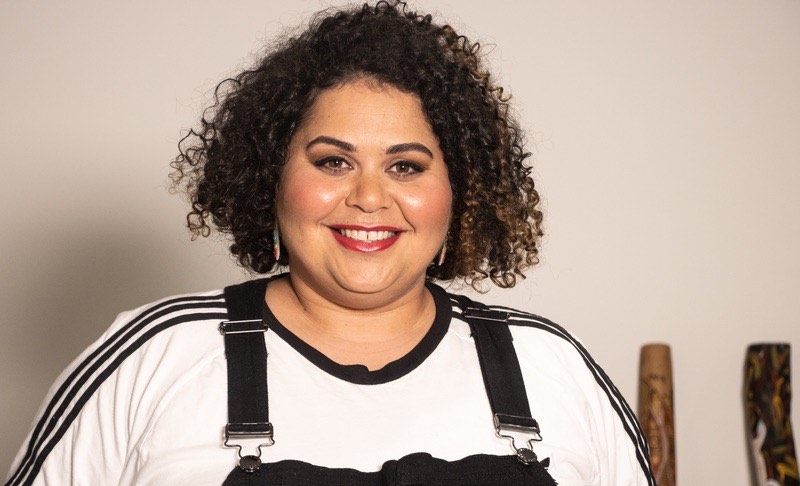 A Headshot of Tarneen Onus Williams. Her chest, shoulders, face and head are shown. She has dark brown hair, that is made up of very tight curls, and reaches her jawline. She is wearing a white t-shirt with the three adidas stripes running down her shoulders, underneath black denim overalls. She has some eyeshadow, blush and deep berry lipstick. She is smiling at the camera.